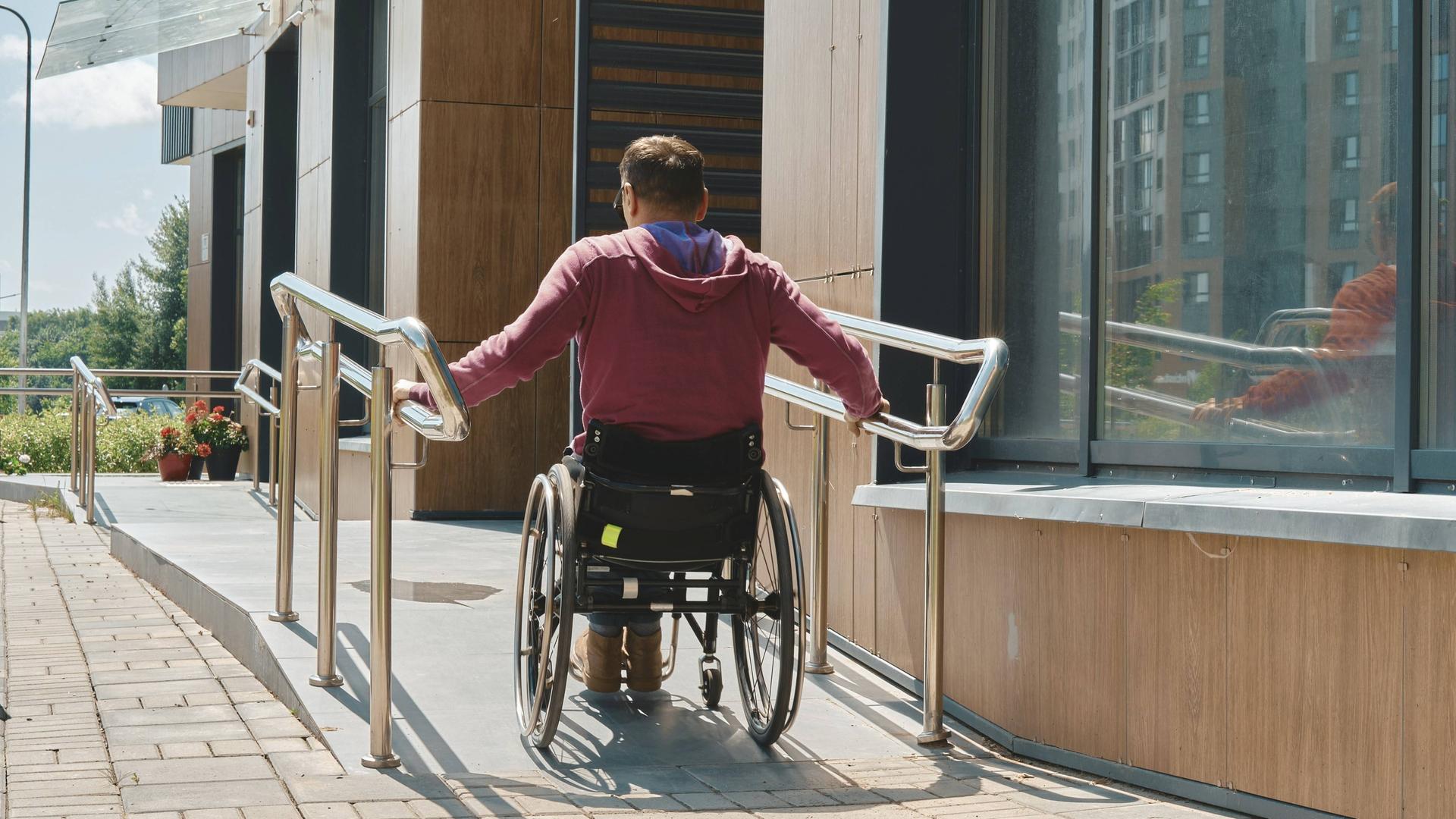 Ein Mann in einem Roll-Stuhl fährt eine Rampe zu dem Eingang an einem Gebäude hoch.