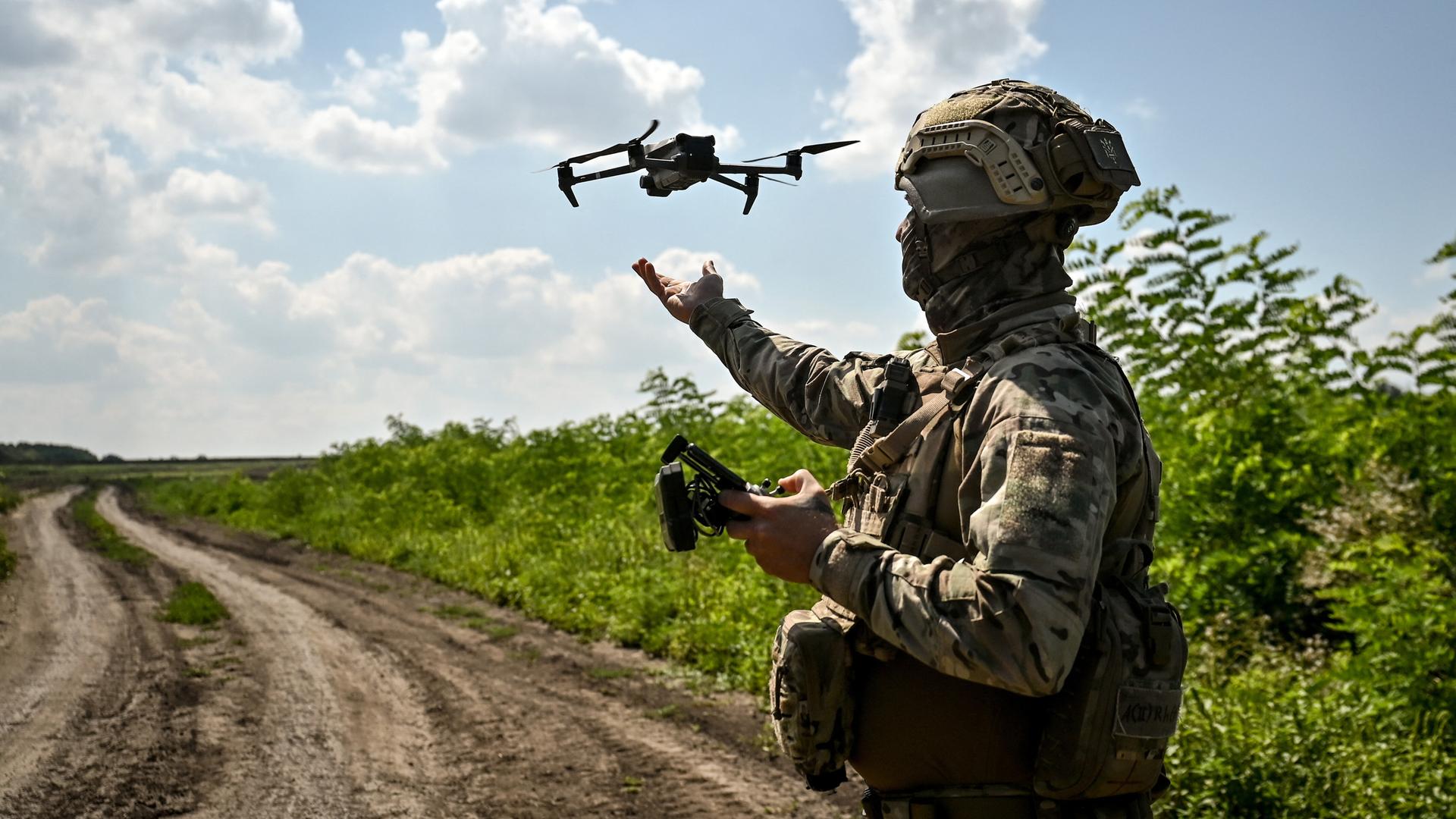Ein Soldat steht auf einem Feldweg. In der Hand hält er eine Fernbedienung, in der Luft fliegt eine Drohne.