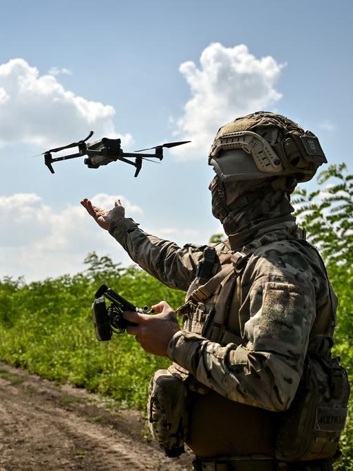 Ein Soldat steht auf einem Feldweg. In der Hand hält er eine Fernbedienung, in der Luft fliegt eine Drohne.