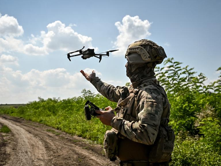 Ein Soldat steht auf einem Feldweg. In der Hand hält er eine Fernbedienung, in der Luft fliegt eine Drohne.