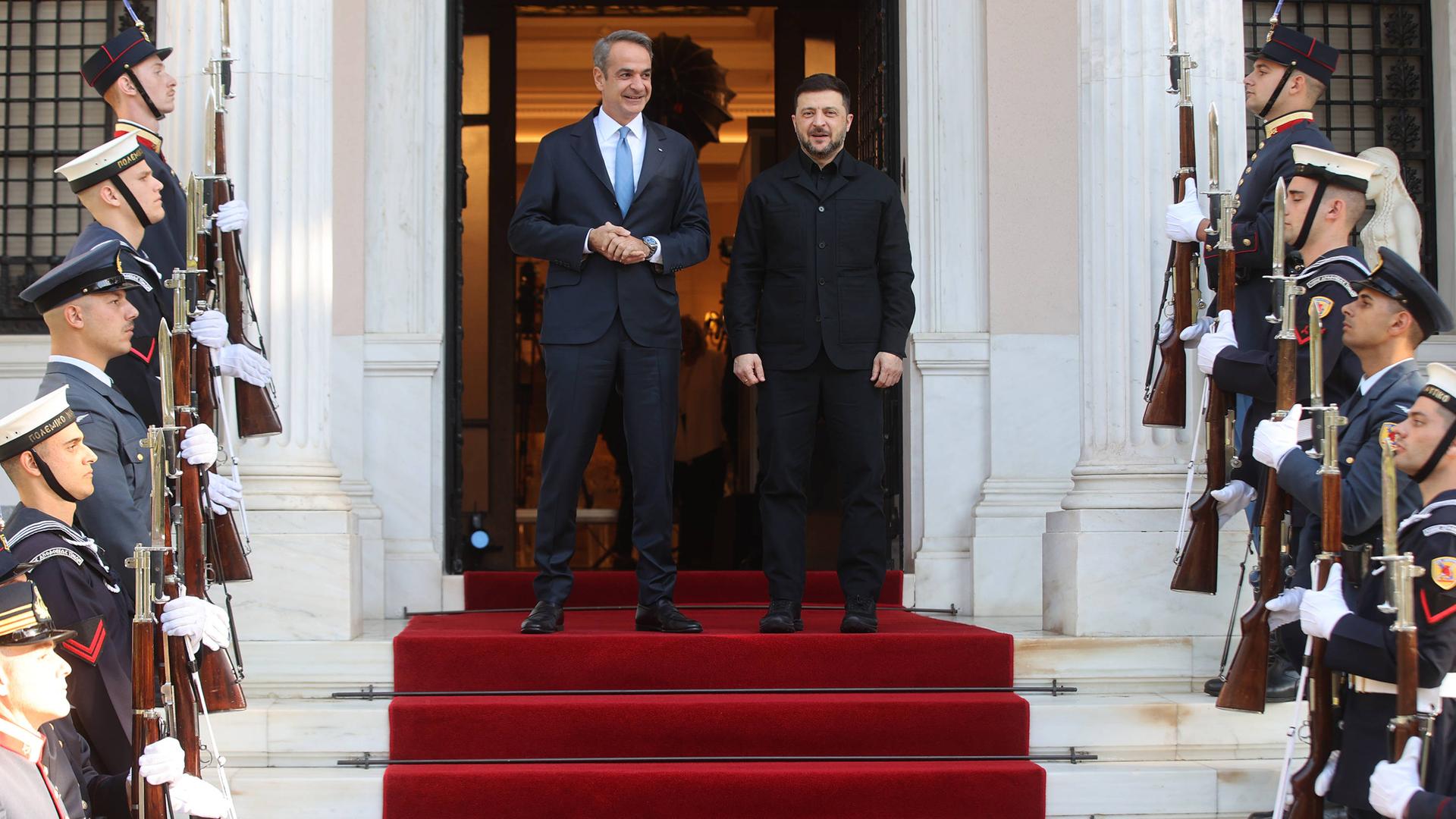 Der ukrainische Präsident Selenskyj wird vom griechischen Regierungschef Mitsotakis empfangen. Die beiden Politiker stehen auf einer Treppe, an den Treppenstufen stehen Soldaten mit Gewehren Spalier. 