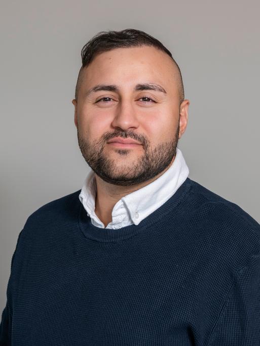 Porträt des Arztes und Autors Dr. Mahmut Martin Yüksel. Er trägt einen blauen Pullover, am Hals schaut ein weißer Hemdkragen hervor. Er blickt selbstbewusst in die Kamera.