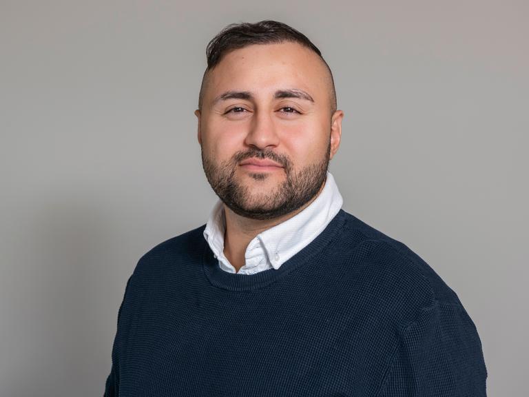 Porträt des Arztes und Autors Dr. Mahmut Martin Yüksel. Er trägt einen blauen Pullover, am Hals schaut ein weißer Hemdkragen hervor. Er blickt selbstbewusst in die Kamera.