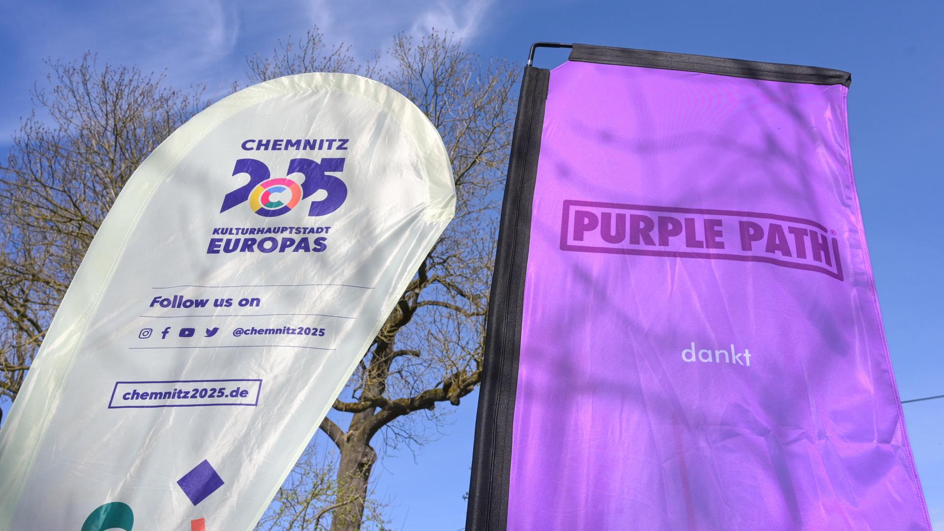 Zwei Flaggen mit der Aufschrift "Chemnitz 2025, Kulturhauptstadt Europas" und "Purple Phat" stehen nebeneinander. Der Kunst- und Skulpturenweg Purple Path verbindet die Orte in der Kulturhauptstadt und Region Chemnitz.