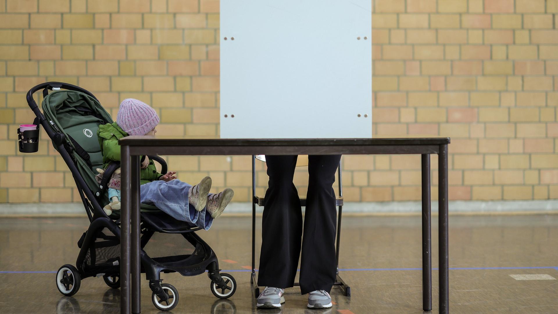 Eine Frau steht in einer Turnhalle in Duisburg in einer Wahl-Kabine. Neben ihr steht ein Kinder-Wagen mit einem Kind. 