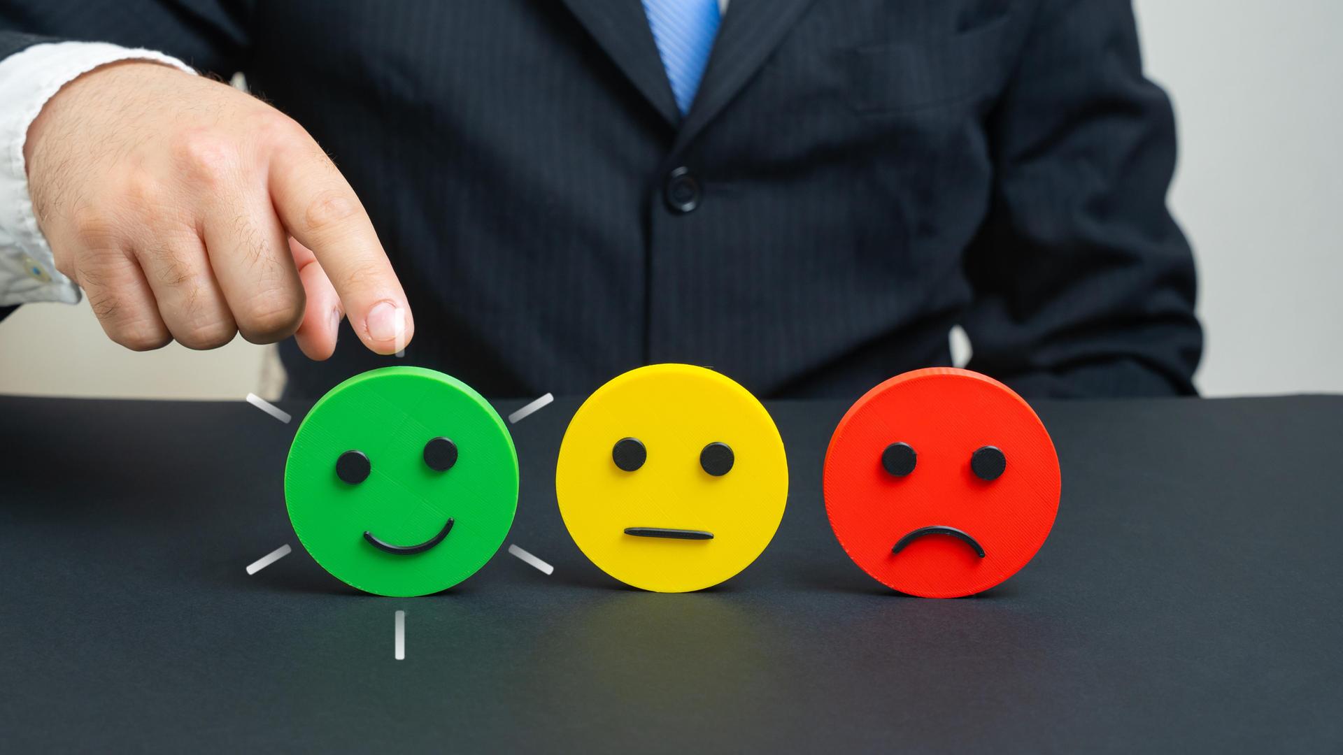 Eine Hand zeigt auf einen grünen Smiley, der lächelt. Daneben steht ein gelber Smiley mit neutralem Ausdruck und ein roter Smiley, der Ablehnung signalisiert.