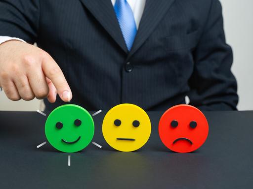 Eine Hand zeigt auf einen grünen Smiley, der lächelt. Daneben steht ein gelber Smiley mit neutralem Ausdruck und ein roter Smiley, der Ablehnung signalisiert. Eine Hand zeigt auf einen grünen Smiley, der lächelt. Daneben steht ein gelber Smiley mit neutralem Ausdruck und ein roter Smiley, der Ablehnung signalisiert.