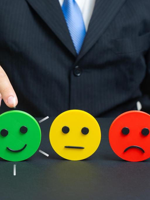 Eine Hand zeigt auf einen grünen Smiley, der lächelt. Daneben steht ein gelber Smiley mit neutralem Ausdruck und ein roter Smiley, der Ablehnung signalisiert. Eine Hand zeigt auf einen grünen Smiley, der lächelt. Daneben steht ein gelber Smiley mit neutralem Ausdruck und ein roter Smiley, der Ablehnung signalisiert.