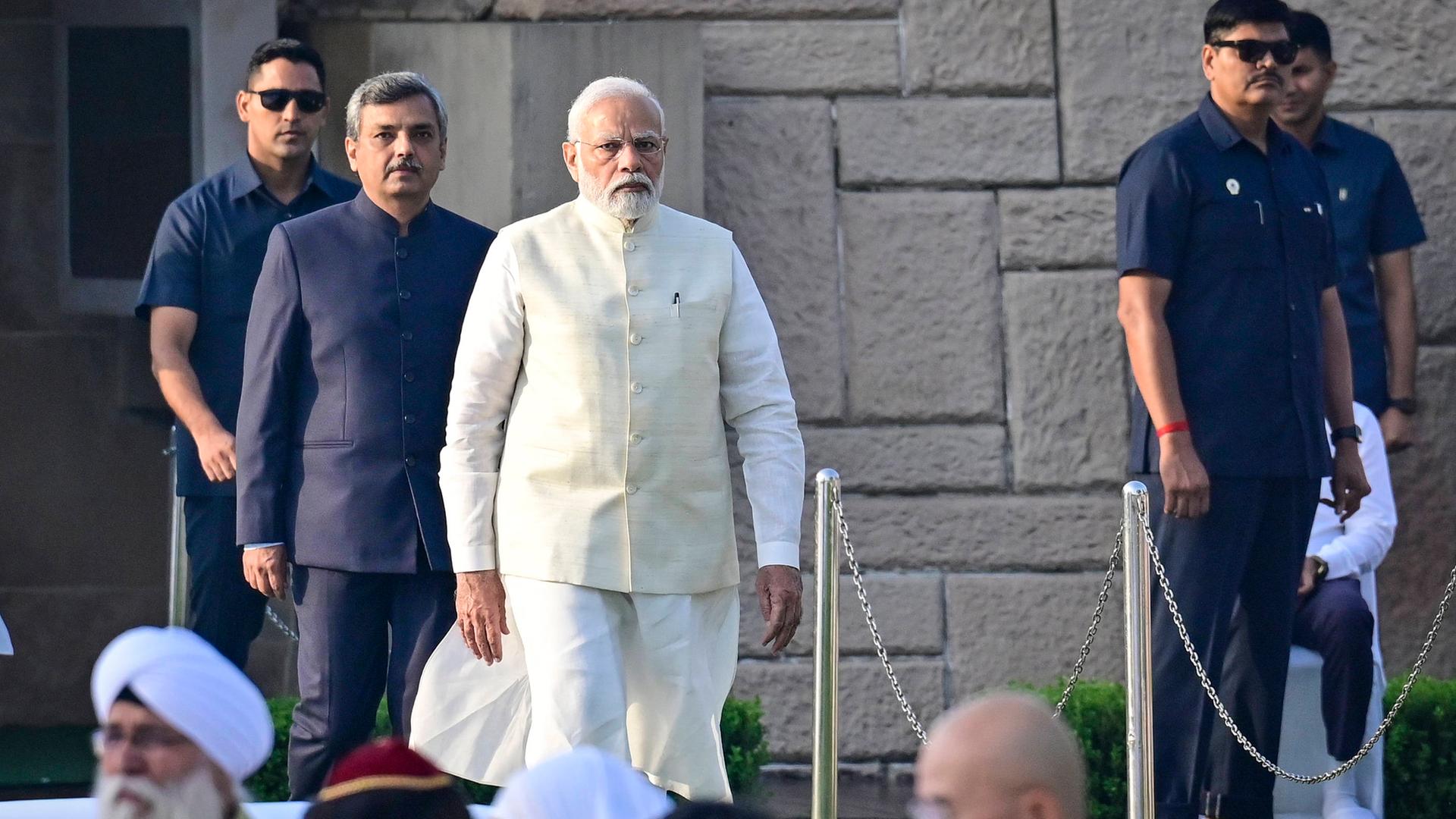 Der indische Premierminister Narendra Modi schreitet in einem weißem Gewand einen markierten Weg ab. Hinter stehen ihm mehrere Sicherheitskräfte.