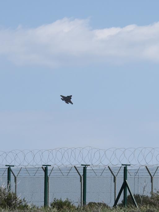 Ein Militärflugzeug startet über dem Meer von Zypern. Im Vordergrund steht ein Zaun mit NATO-Draht.