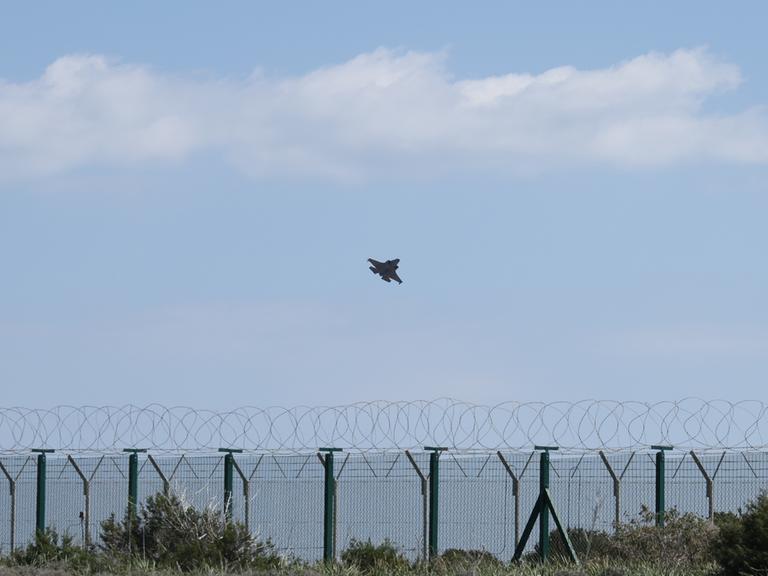 Ein Militärflugzeug startet über dem Meer von Zypern. Im Vordergrund steht ein Zaun mit NATO-Draht.