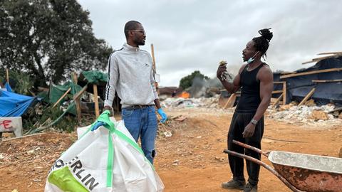 Zwei Persons of Color stehen sich im Gespräch gegenüber. Der linke trägt eine große Schutttüte, der rechte steht an einer Schubkarre.
