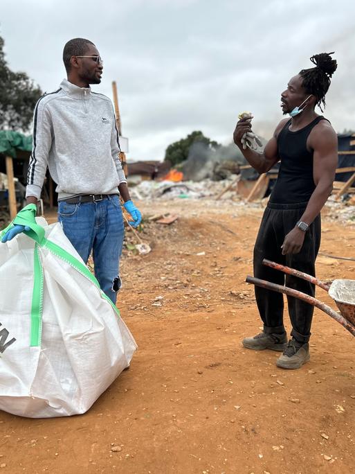 Zwei Persons of Color stehen sich im Gespräch gegenüber. Der linke trägt eine große Schutttüte, der rechte steht an einer Schubkarre.