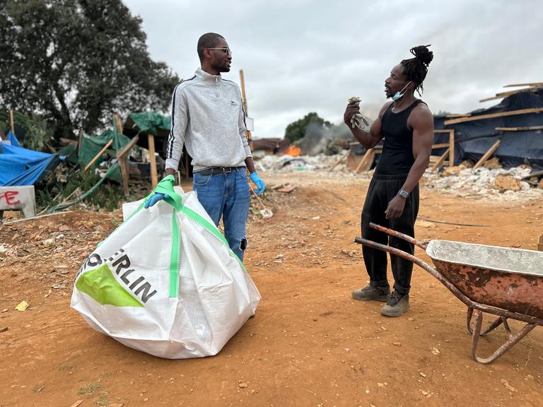 Zwei Persons of Color stehen sich im Gespräch gegenüber. Der linke trägt eine große Schutttüte, der rechte steht an einer Schubkarre.