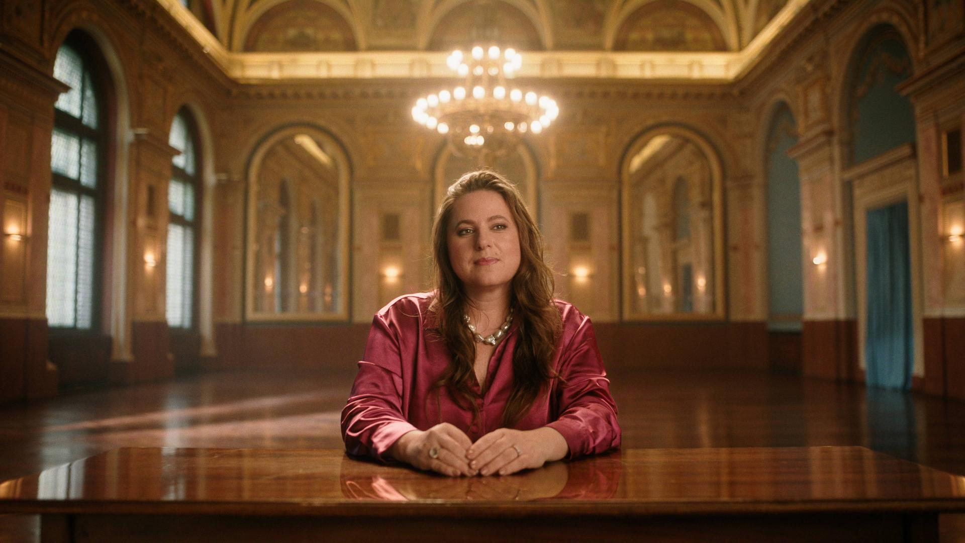 Judit Polgar sitzt an einem großen Tisch und schaut in die Kamera. Hinter ihr ist ein prunkvoller Saal zu sehen. 