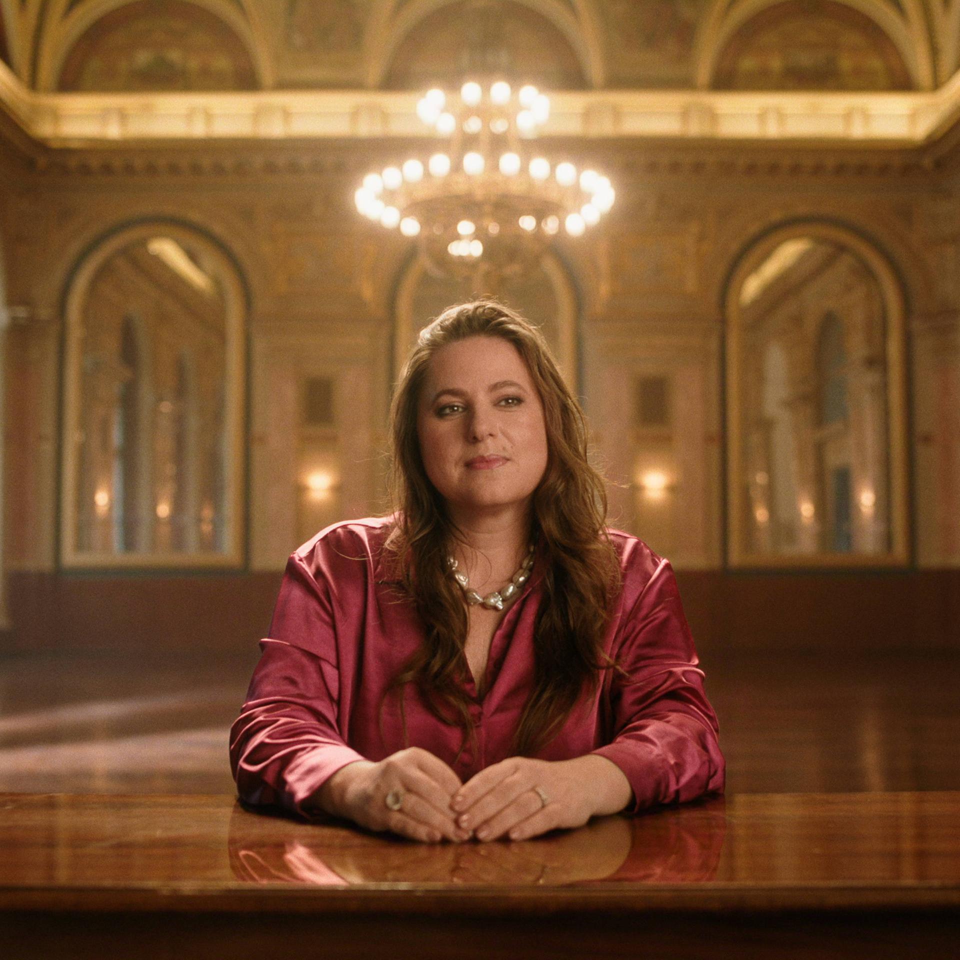 Judit Polgar sitzt an einem großen Tisch und schaut in die Kamera. Hinter ihr ist ein prunkvoller Saal zu sehen. 