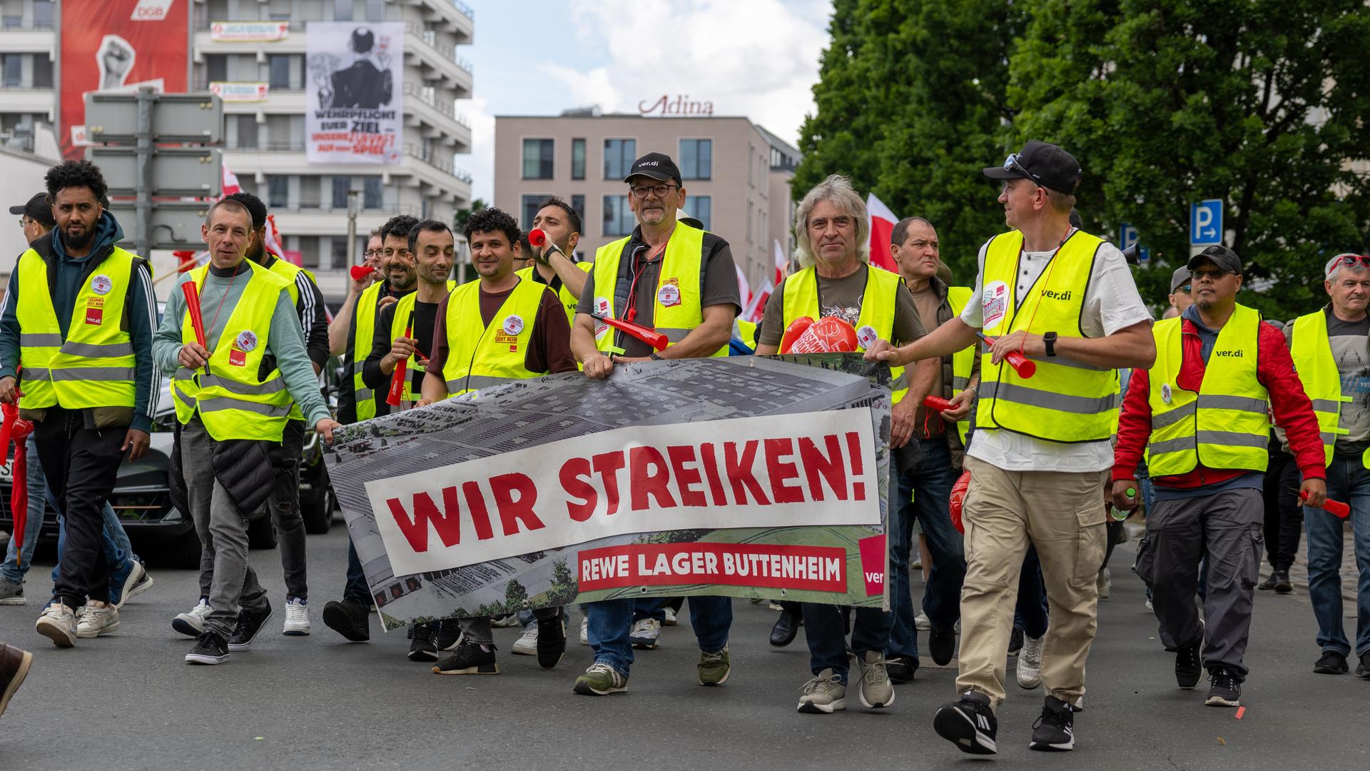 Die Streikenden tragen gelbe Warnwesten und laufen nebeneinander eine Straße entlang. Mehrere von ihnen halten ein Transparent mit der Aufschrift "Wir streiken! REWE Lager Buttenheim" vor sich. 
