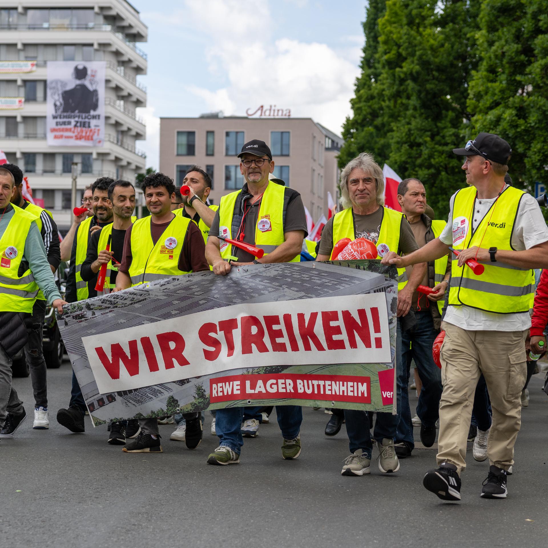 Die Streikenden tragen gelbe Warnwesten und laufen nebeneinander eine Straße entlang. Mehrere von ihnne halten ein Transparent mit der Aufschrift "Wir streiken! REWE Lager Buttenheim" vor sich. 