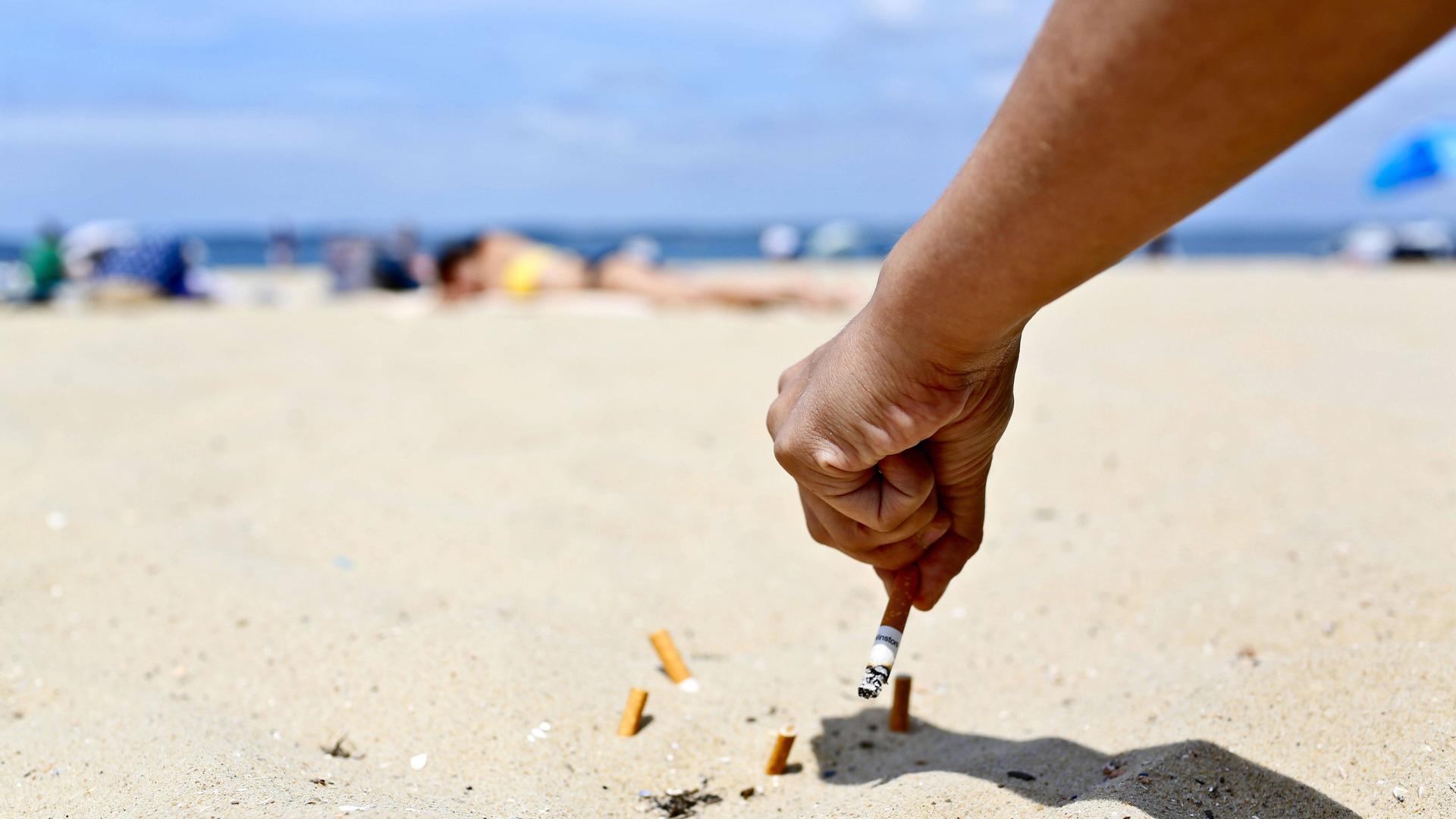 An einem sonnigen Sandstrand hält eine Hand eine Zigarette und drückt diese im Sand aus. Dort liegen bereits mehrere Zigarettenstummel. An einem sonnigen Sandstrand hält eine Hand eine Zigarette und drückt diese im Sand aus. Dort liegen bereits mehrere Zigarettenstummel.