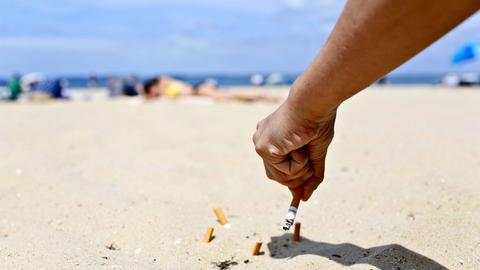 An einem sonnigen Sandstrand hält eine Hand eine Zigarette und drückt diese im Sand aus. Dort liegen bereits mehrere Zigarettenstummel.