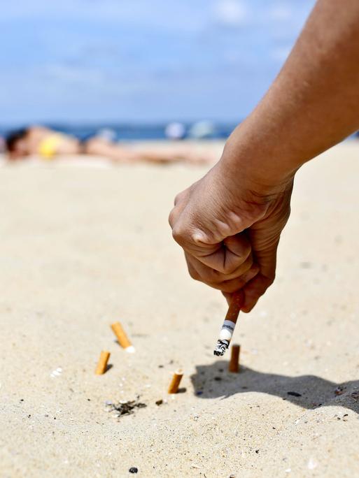 An einem sonnigen Sandstrand hält eine Hand eine Zigarette und drückt diese im Sand aus. Dort liegen bereits mehrere Zigarettenstummel.