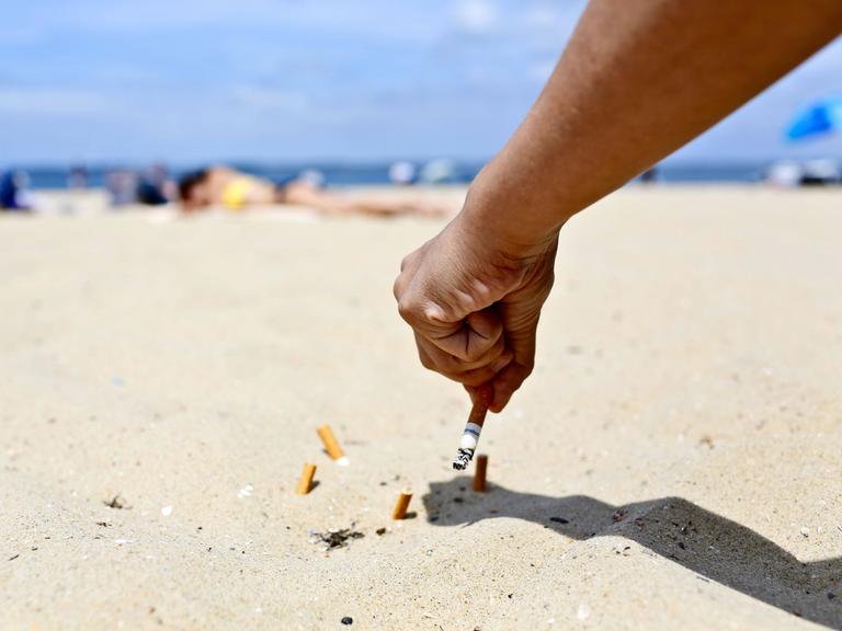 An einem sonnigen Sandstrand hält eine Hand eine Zigarette und drückt diese im Sand aus. Dort liegen bereits mehrere Zigarettenstummel.
