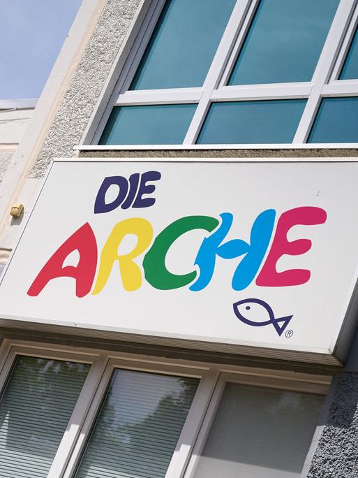An einer Hauswand hängt ein Schild mit der Aufschrift Die Arche in bunten Buchstaben, darunter ist ein Fisch gemalt. Links vom Schild hängt ein Banner mit der Aufschrift Lasst die Kinder zu mir kommen und hindert sie nicht daran, Die Bibel.