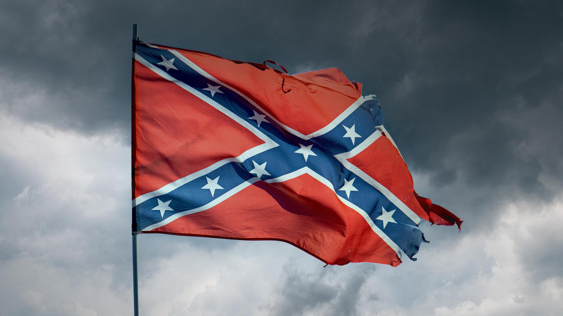 Eine im Wind wehende, zerfledderte "Rebellenflagge", ein weißumrahmtes blaues Schragenkreuz bedeckt mit dreizehn weißen Sternen auf rotem Grund, vor einem grau bewölkten Himmel mit aufziehendem Gewitter. 