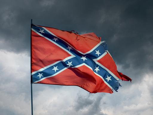 Eine im Wind wehende, zerfledderte "Rebellenflagge", ein weißumrahmtes blaues Schragenkreuz bedeckt mit dreizehn weißen Sternen auf rotem Grund, vor einem grau bewölkten Himmel mit aufziehendem Gewitter. 