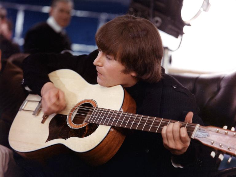 John Lennon sitzt auf einem Sessel und spielt eine Gitarre. 