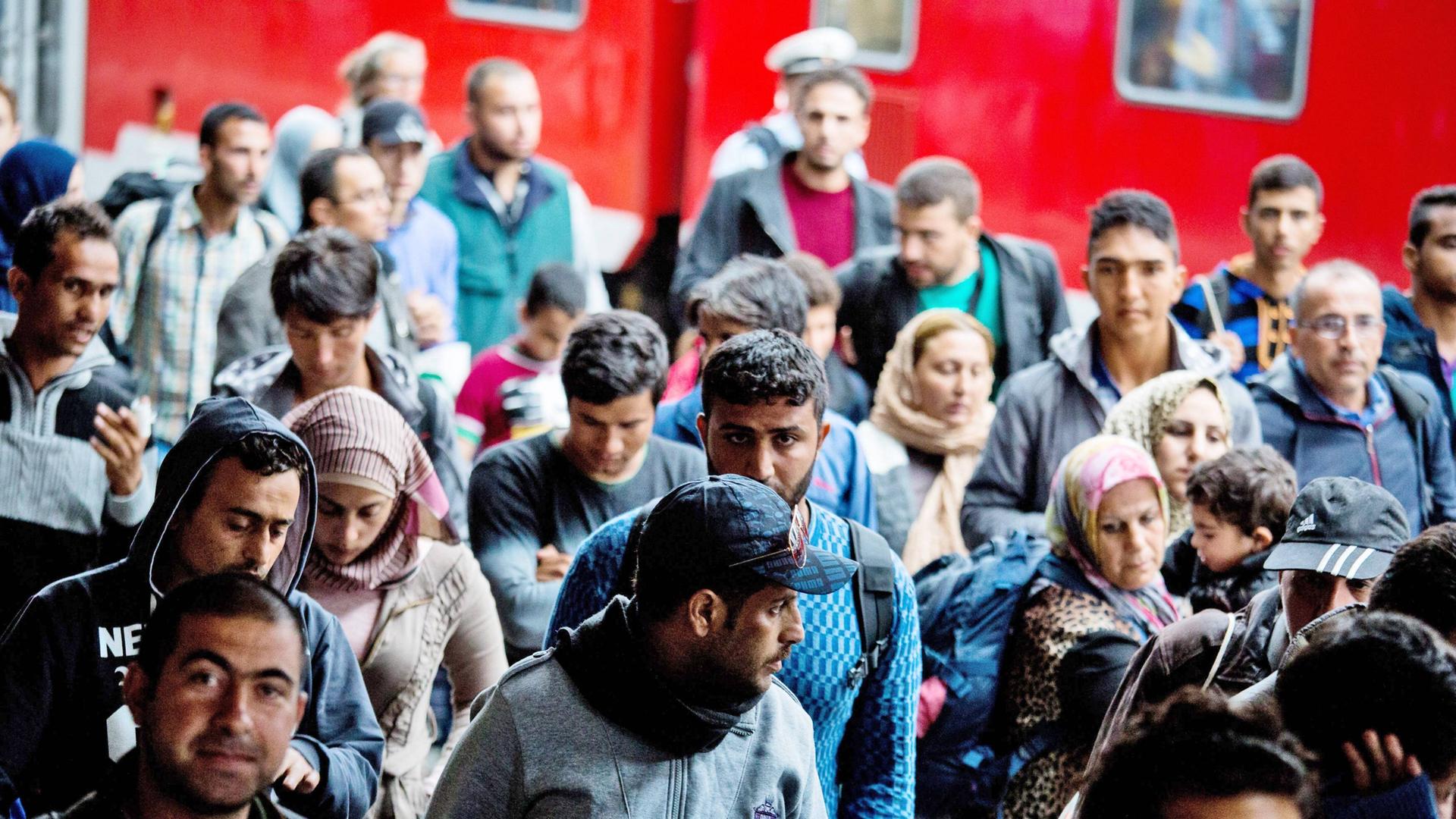 Das Bild zeigt eine Gruppe von Flüchtlingen Im Hintergrund ist ein roter Zugwaggon zu sehen.