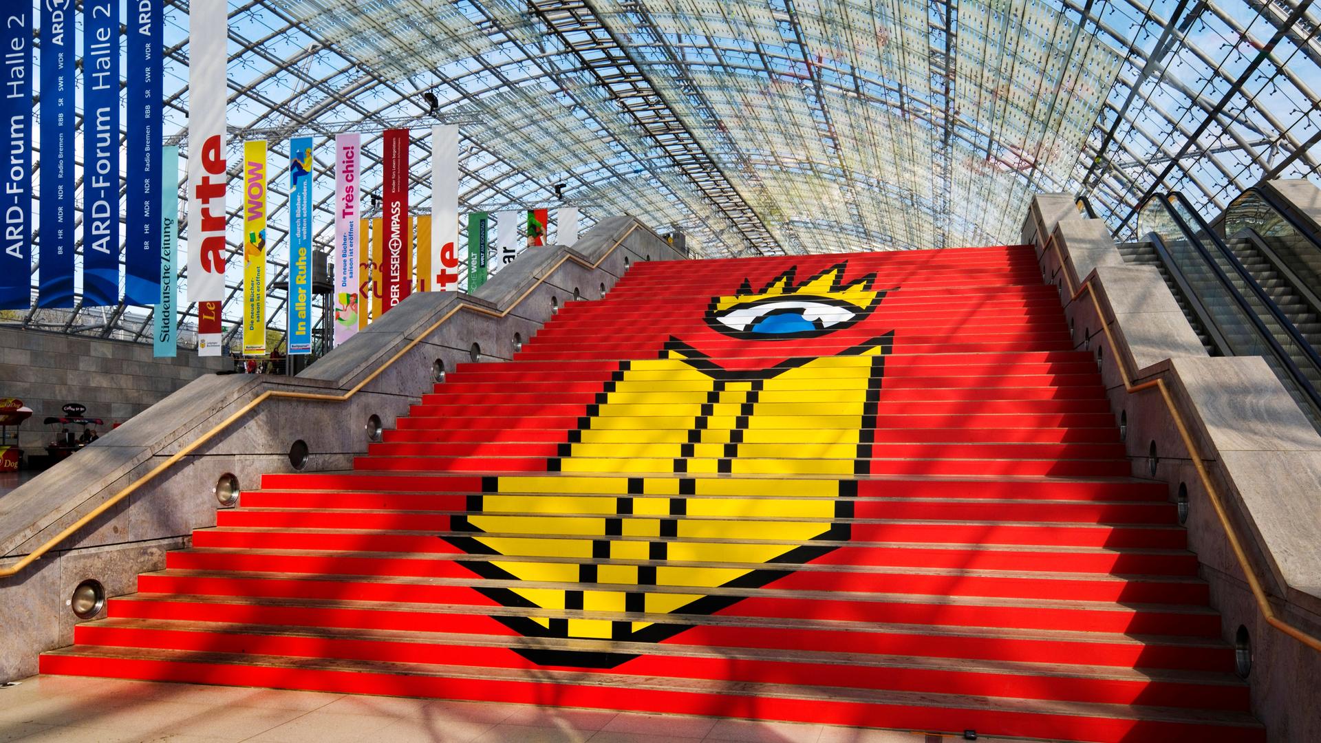 Das Logo der Leipziger Buchmesse auf einer Treppe in einer Glashalle der Messe in Leipzig. Auf rotem Grund ist ein stilisierter Kopf zu sehen, der aus nur einem Auge besteht. Davor, ebenfalls in Gelb, die Umrisse eines Buches. 