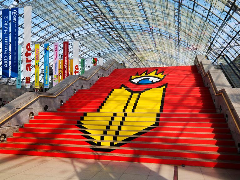 Das Logo der Leipziger Buchmesse auf einer Treppe in einer Glashalle der Messe in Leipzig. Auf rotem Grund ist ein stilisierter Kopf zu sehen, der aus nur einem Auge besteht. Davor, ebenfalls in Gelb, die Umrisse eines Buches. 