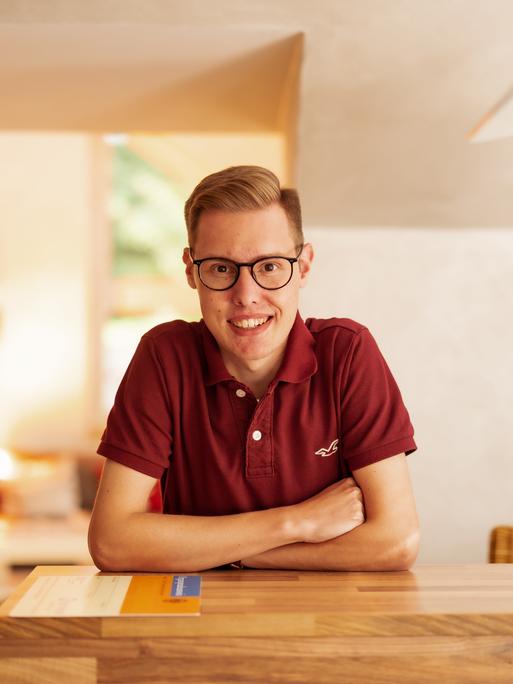 Porträt von Marius Schaefer aus Arnsberg-Oeventrop, der als erster Mensch in Deutschland eine Lebendlungenspende von seinen Eltern erhielt. Er stützt sich mit verschrenkten Armen auf einen Tisch und blickt freundich in die Kamera