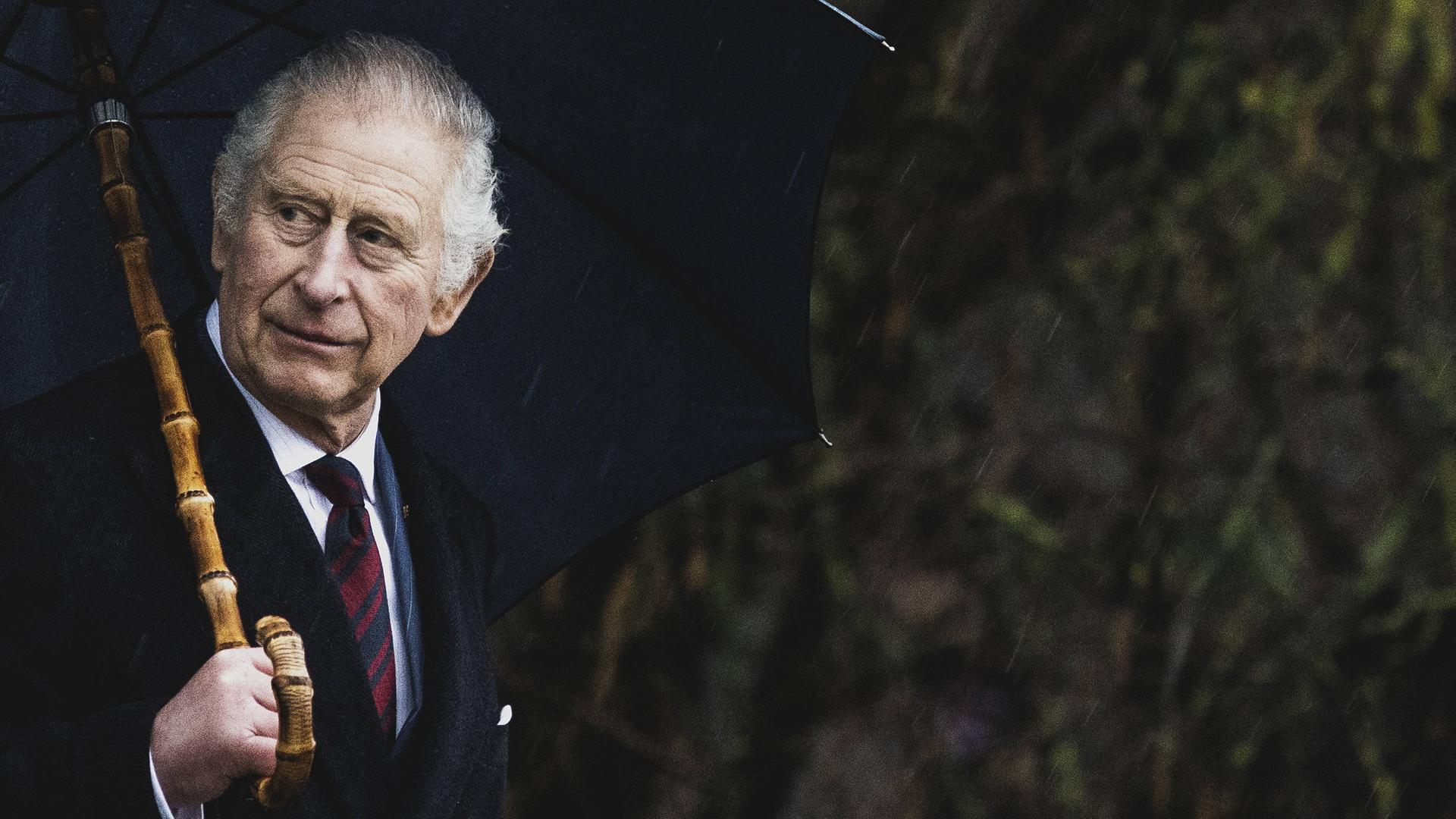 König Charles der III. vor dunklem Hintergrund. In der linken Hand hält er einen Regenschirm und guckt links aus dem Bild raus.