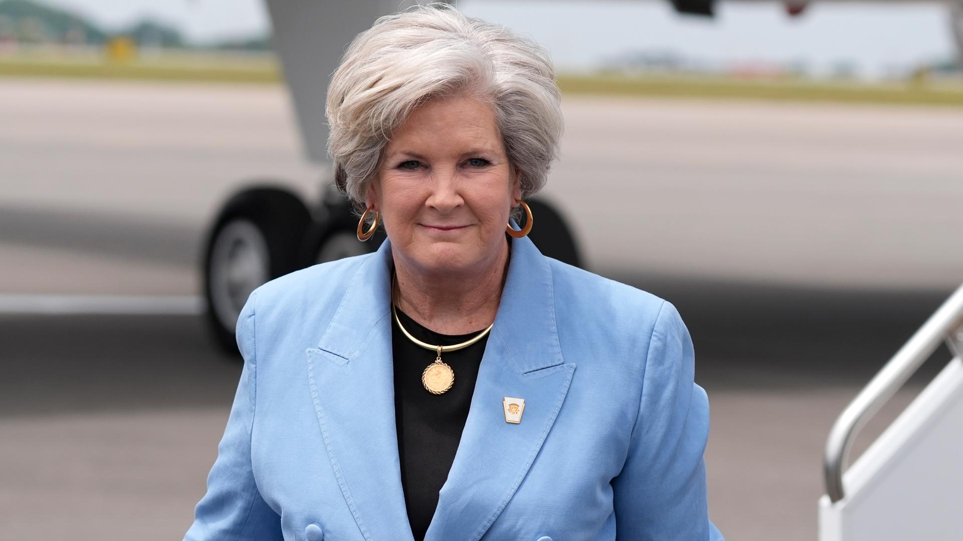 Das Bild zeigt Susan Wiles vor einem Flugzeug. Sie trägt einen blauen Blazer und schaut in die Kamera.