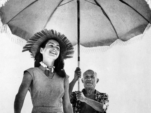 Pablo Picasso läuft am Strand von Cannes hinter Françoise Gilot und hält einen Sonnenschirm in den Händen.