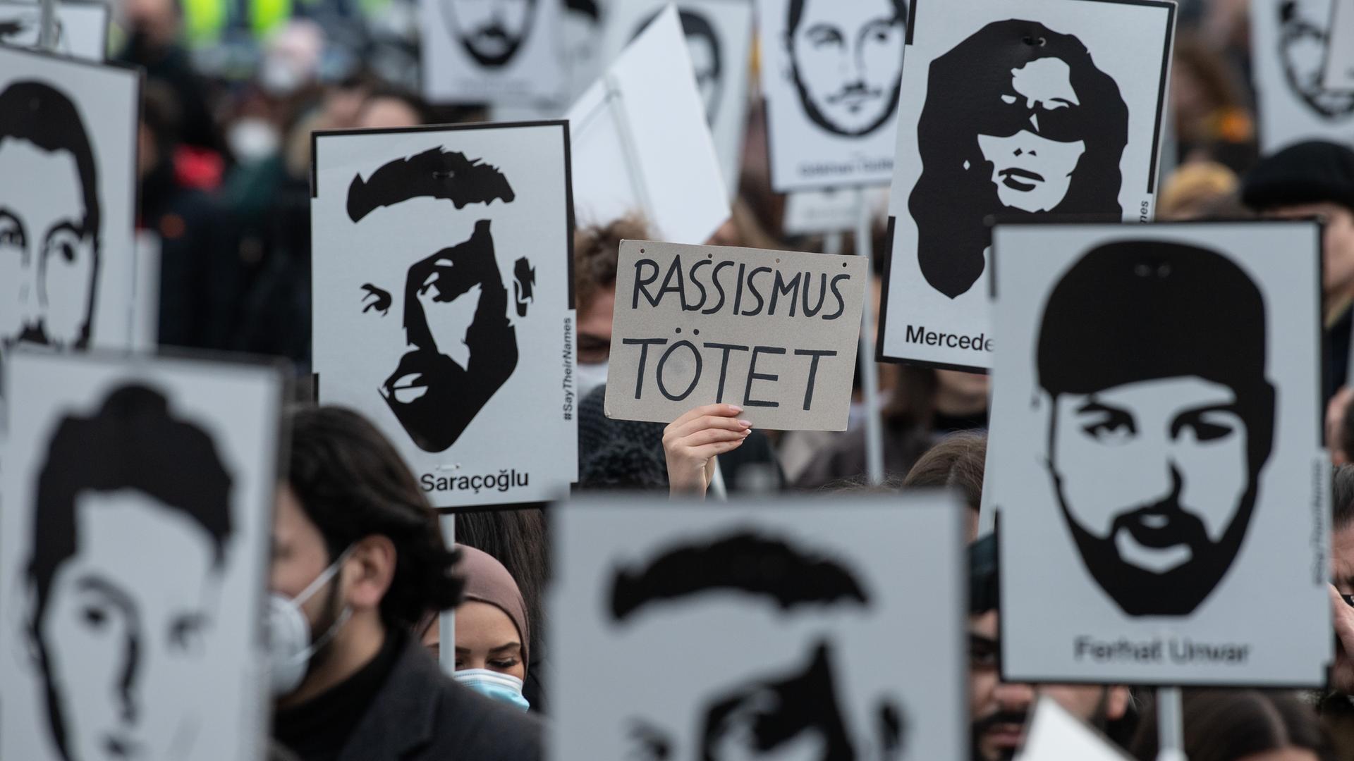 Mit Plakaten und Bildern der Ermordeten erinnern Teilnehmer einer Gedenkveranstaltung auf dem Marktplatz von Hanau an die Opfer der rassistisch motivierten Anschläge von Hanau im Jahr 2020. Auf einem Schild steht "Rassismus tötet".