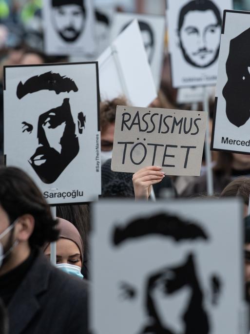 Mit Plakaten und Bildern der Ermordeten erinnern Teilnehmer einer Gedenkveranstaltung auf dem Marktplatz von Hanau an die Opfer der rassistisch motivierten Anschläge von Hanau im Jahr 2020. Auf einem Schild steht "Rassismus tötet".