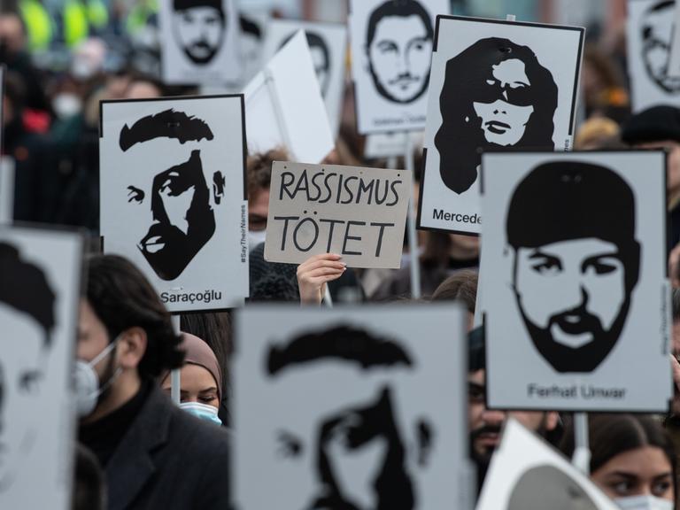 Mit Plakaten und Bildern der Ermordeten erinnern Teilnehmer einer Gedenkveranstaltung auf dem Marktplatz von Hanau an die Opfer der rassistisch motivierten Anschläge von Hanau im Jahr 2020. Auf einem Schild steht "Rassismus tötet".