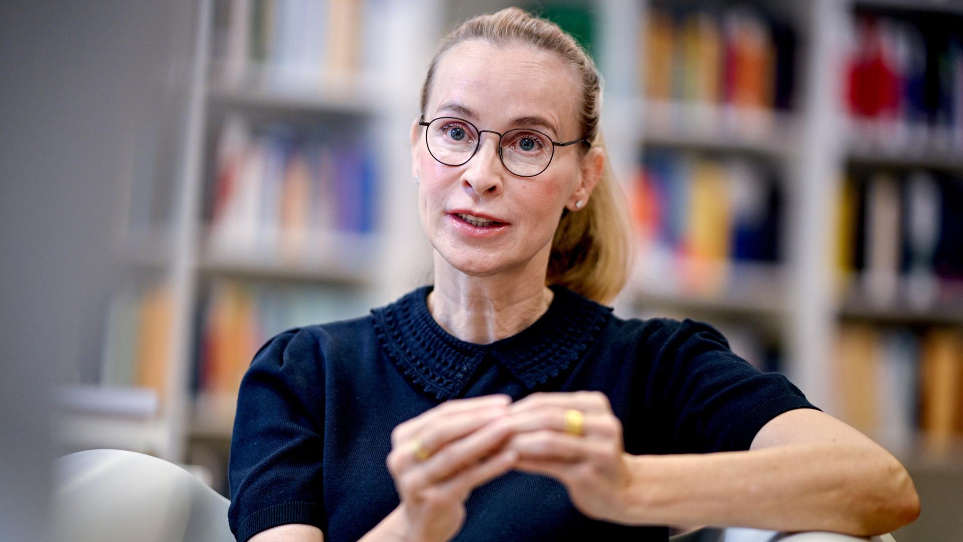 Frauke Brosius-Gersdorf im Porträt. Sie trägt eine dunkle Brille, ein dunkles Oberteil und gestikuliert. Im Hintergrund sind verschwommen Bücherregale zu sehen.