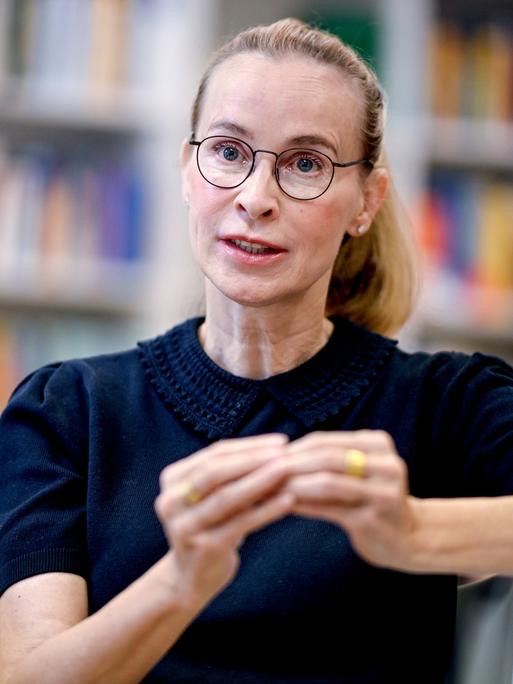 Frauke Brosius-Gersdorf im Porträt. Sie trägt eine dunkle Brille, ein dunkles Oberteil und gestikuliert. Im Hintergrund sind verschwommen Bücherregale zu sehen.