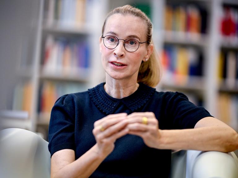 Frauke Brosius-Gersdorf im Porträt. Sie trägt eine dunkle Brille, ein dunkles Oberteil und gestikuliert. Im Hintergrund sind verschwommen Bücherregale zu sehen.