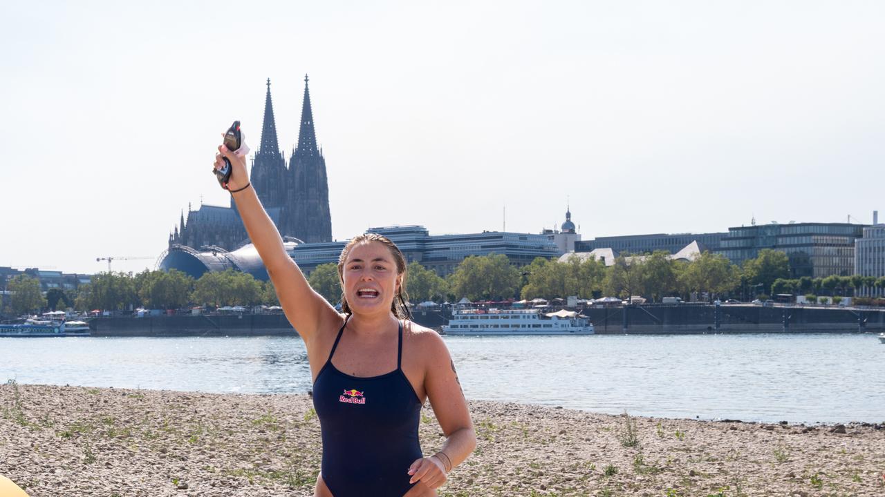 Die Extremschwimmerin Nathalie Pohl jubelt, nachdem sie das Kölner Rheinufer erreicht hat. Sie reißt einen Arm in die Luft. Im Hintergrund ist der Kölner Dom zu sehen. 