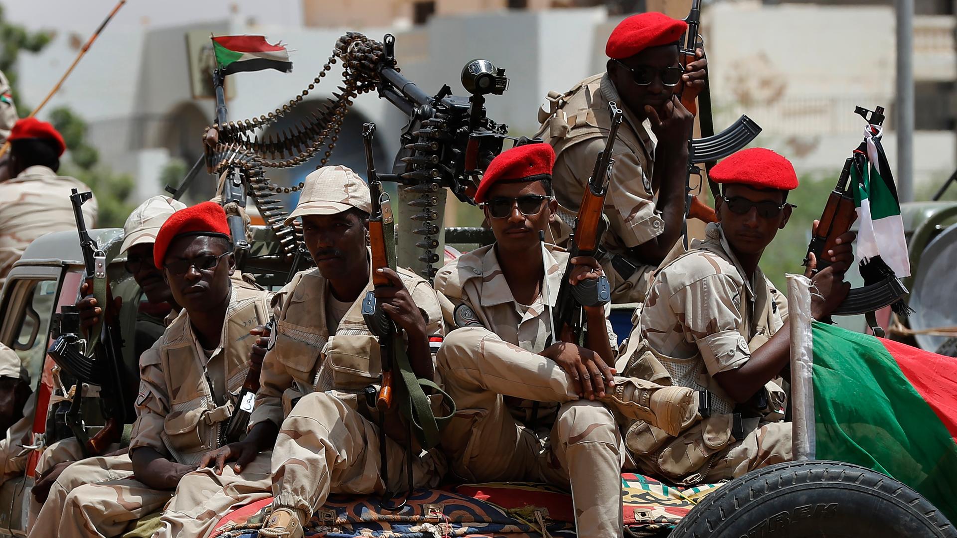 Die Soldaten in Tarnfarben und mit Sonnebrillen, roten Barretten und Gewehren sitzen auf einem Fahrzeug, an dem die Fahne des Sudan hängt. Auf dem Fahrzeug ist ein Maschinengewehr befestigt. Die Soldaten in Tarnfarben und mit Sonnebrillen, roten Barretten und Gewehren sitzen auf einem Fahrzeug, an dem die Fahne des Sudan hängt. Auf dem Fahrzeug ist ein Maschinengewehr befestigt.