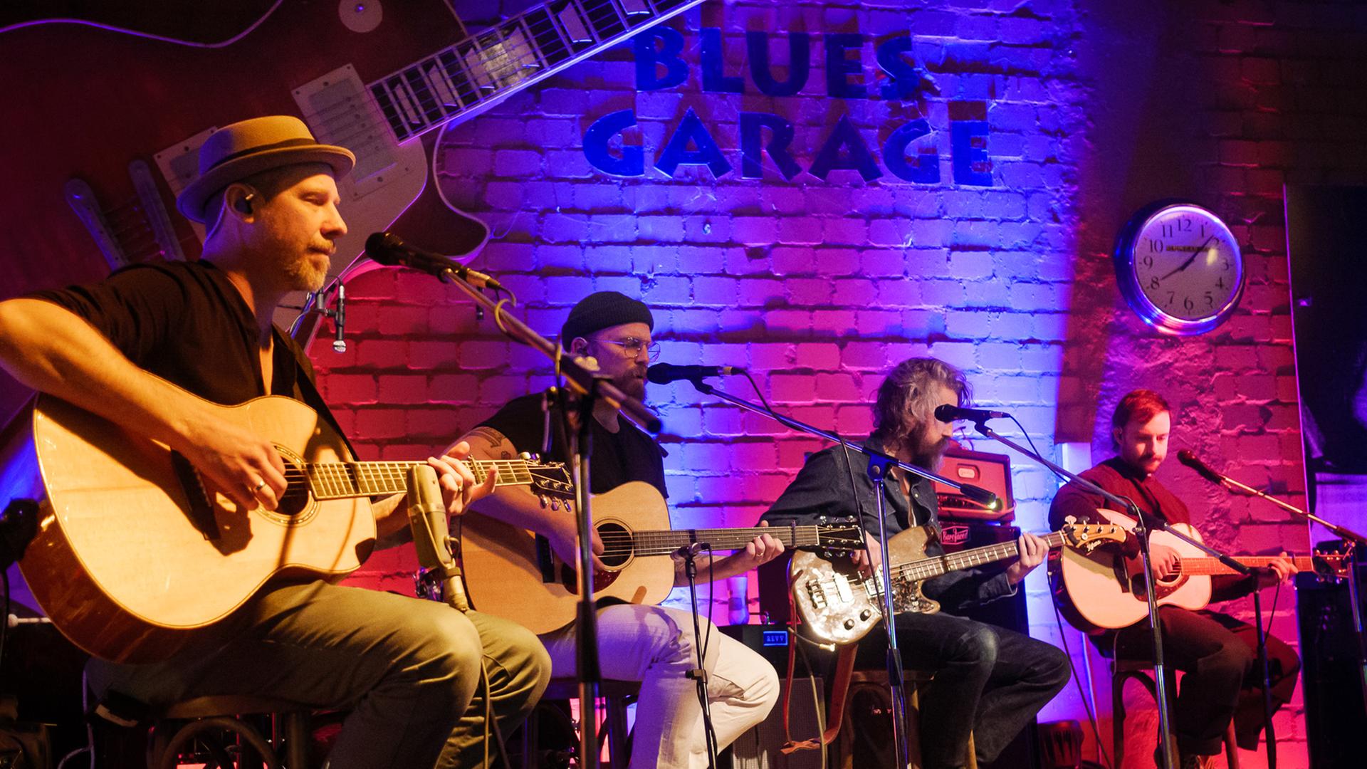 Vier Männer mit Gitarren sitzen auf einer Bühne, spielen und singen.