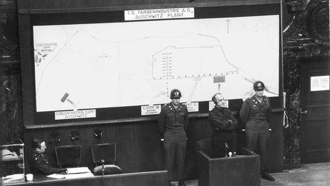 Historisches schwarz-weiß Foto von der Verhandlung im Justizpalast Nürnberg: Oswald Pohl (im Folgeprozess IV gegen die SS zum Tode verurteilt), als Zeuge der Anklage im Zeugenstand, an der Wand ein Lageplan der IG-Farben-Betriebe im KZ-Auschwitz.