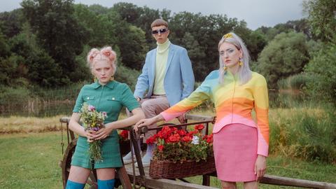 Die dreiköpfige Band Blond, bestehend aus den beiden Schwestern Nina und Lotta Kummer sowie Johann Bonitz posieren auf einer Wiese vor und auf einem blumenbestückten, alten Holzanhänger. Sie tragen poppig-bunte Kleidung.