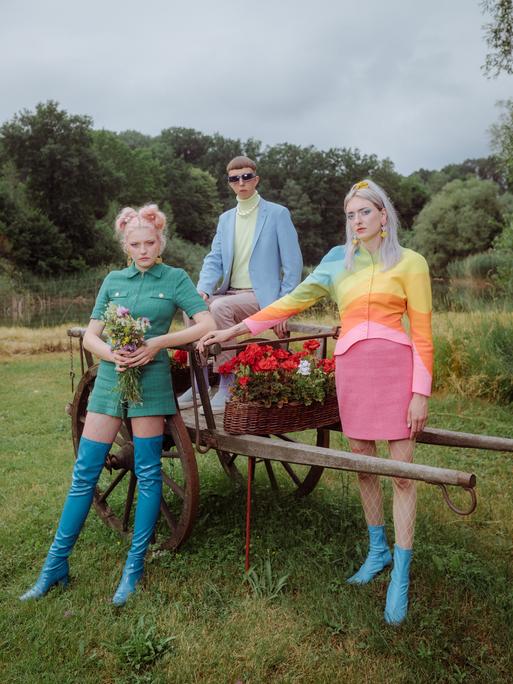 Die dreiköpfige Band Blond, bestehend aus den beiden Schwestern Nina und Lotta Kummer sowie Johann Bonitz posieren auf einer Wiese vor und auf einem blumenbestückten, alten Holzanhänger. Sie tragen poppig-bunte Kleidung.