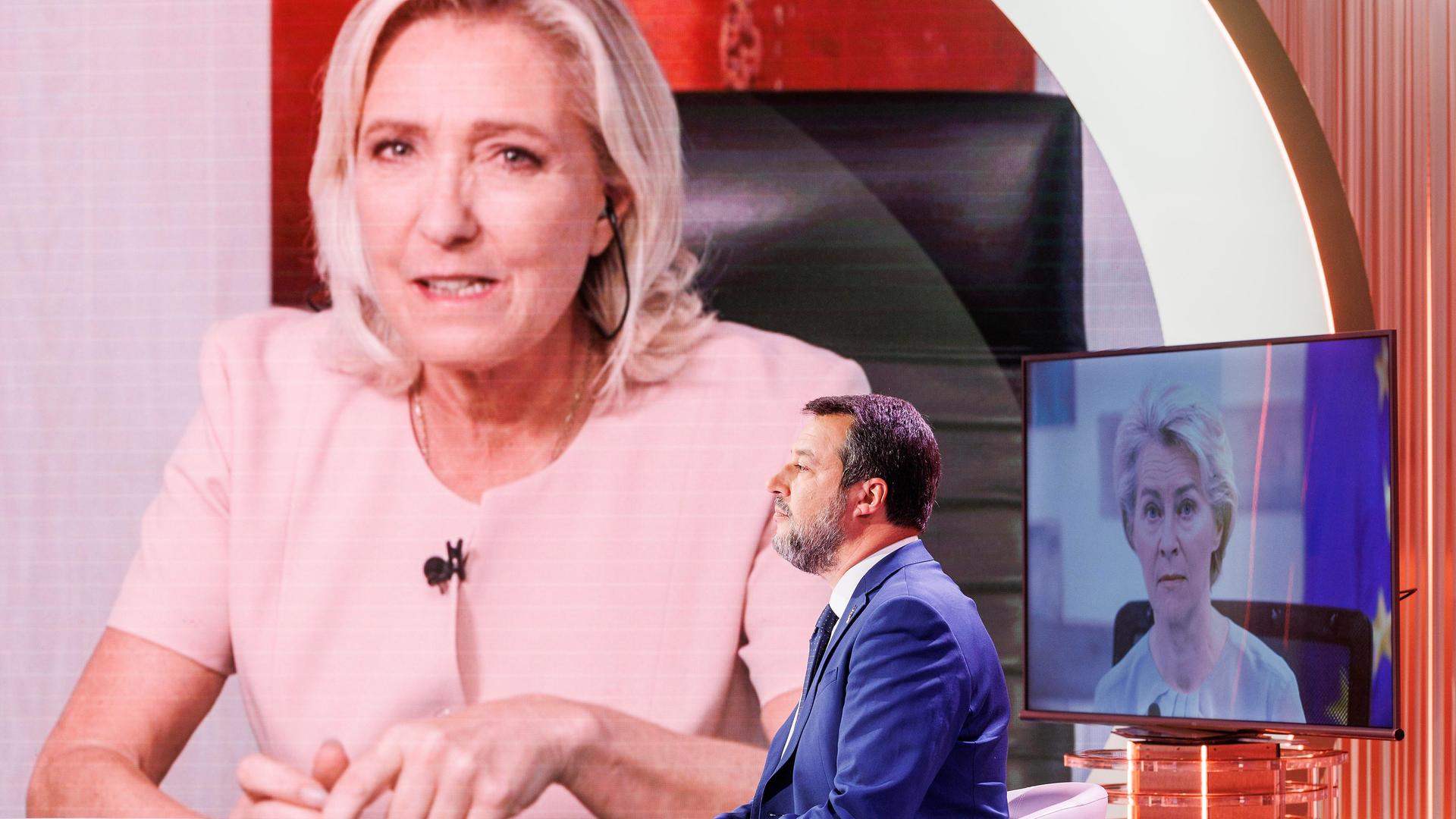 Ein Mann mit blauem Anzug sitzt in einem Fernsehstudio. Neben ihm sind Bildschirme. Darauf sind die Bilder zweier Frauen: Marine Le Pen und Ursula von der Leyen. Der Mann ist der rechte italienische Politiker Matteo Salvini.